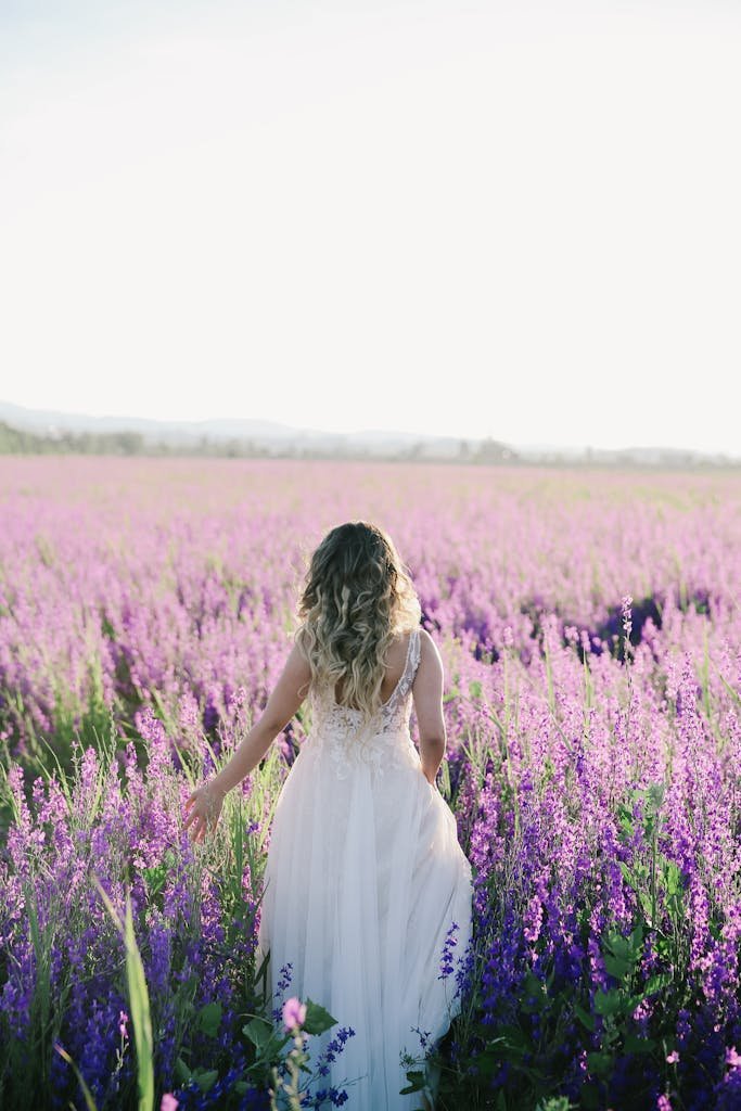 A bride in a white dress walks through a lavender field bathed in sunlight.