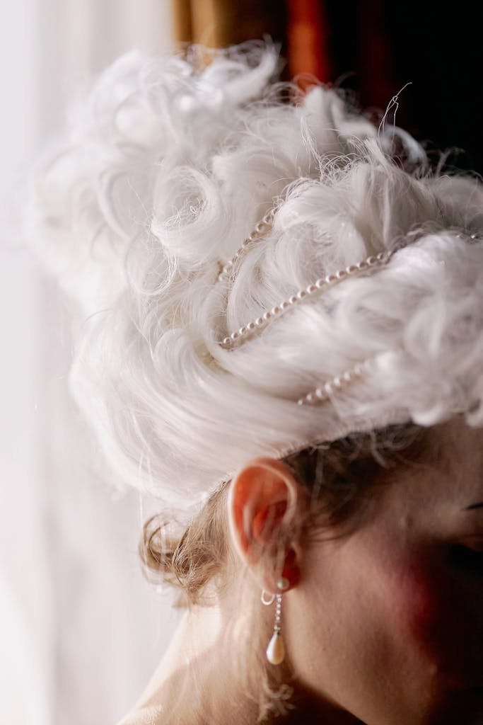 A close-up of an elegant white wig adorned with pearls and an earring, in a vintage style setting.