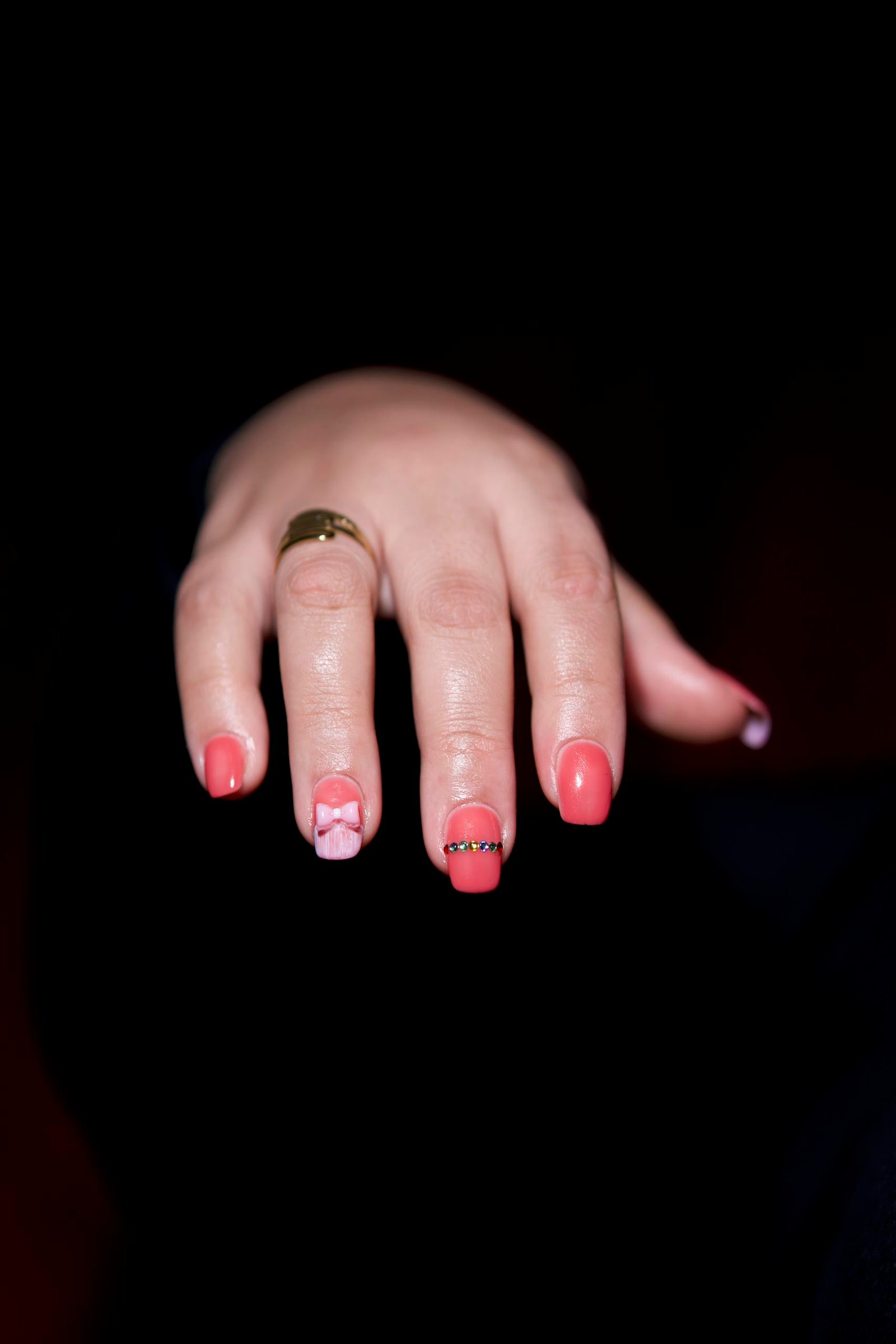 Close-up of a hand with intricate pink nail art and a gold ring against a dark background.