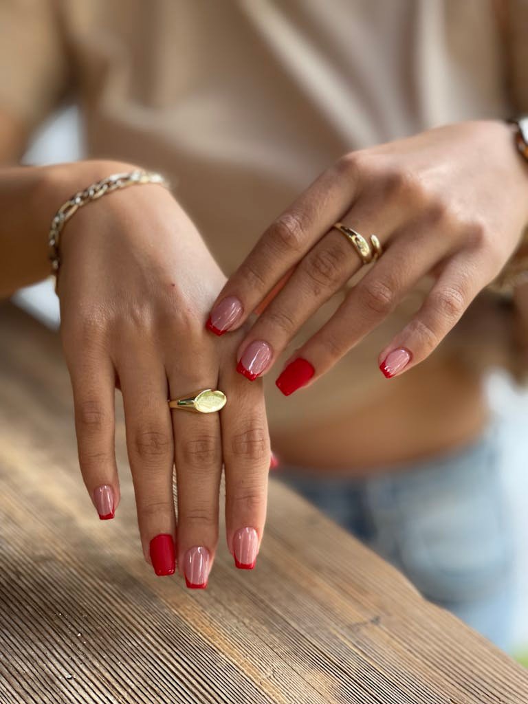 Close-up of beautifully manicured hands with red nail polish and gold rings resting on a wooden surface.