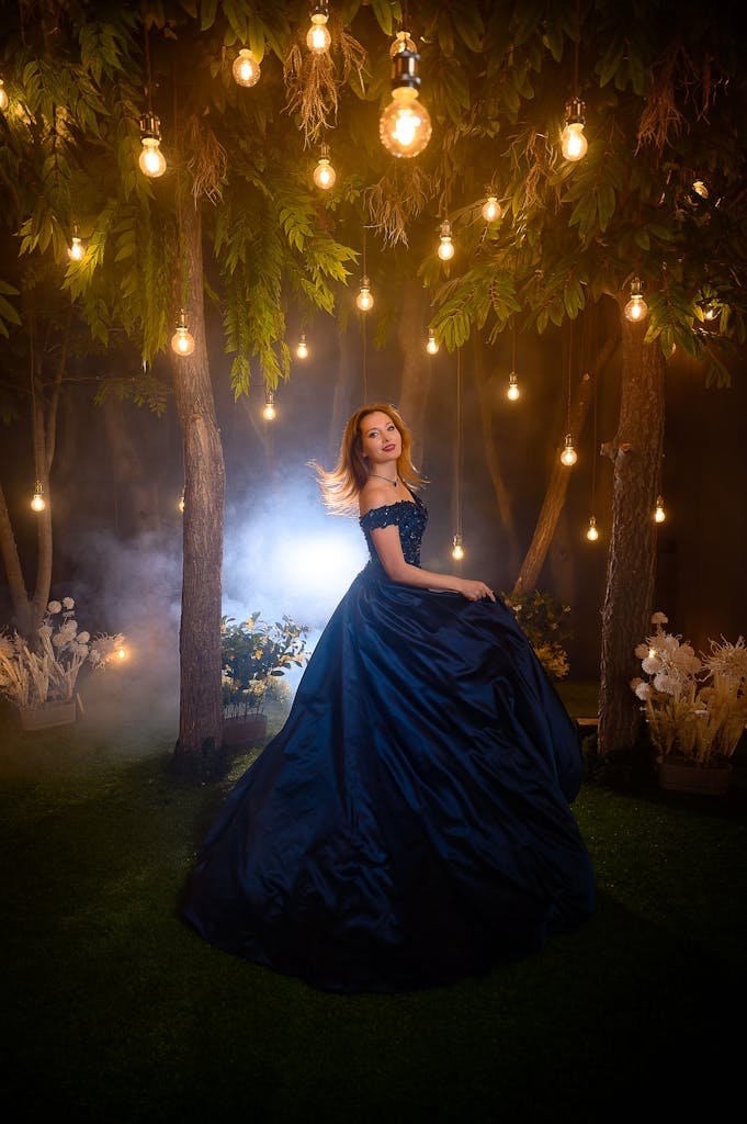 Elegant woman in a navy blue gown posing under hanging lights in an outdoor setting.