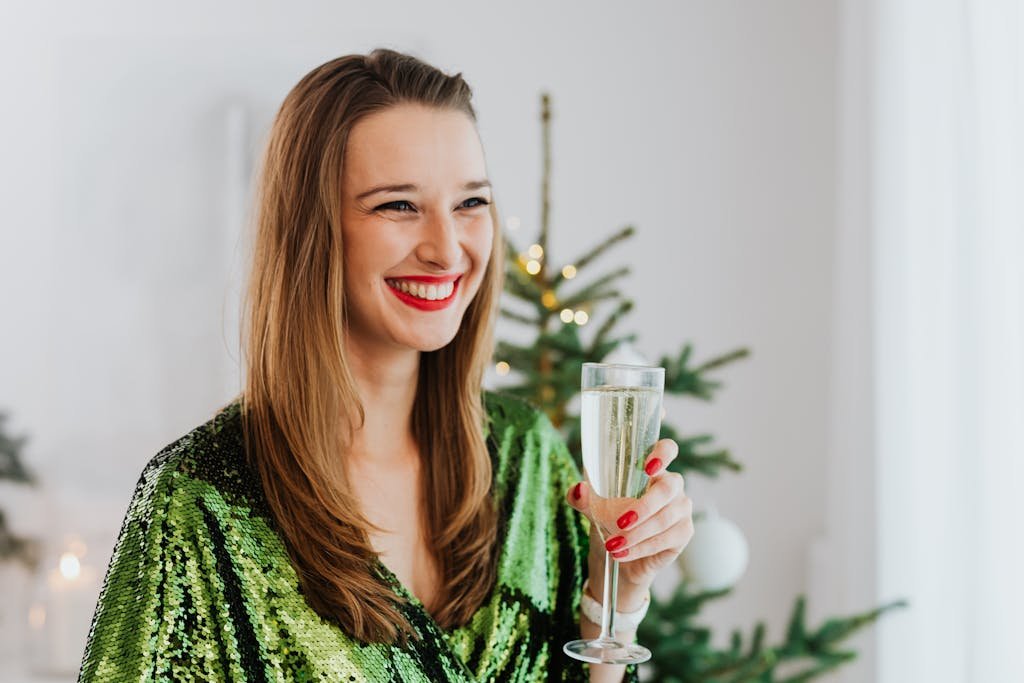 Smiling woman in green dress holding champagne glass by Christmas tree indoors.