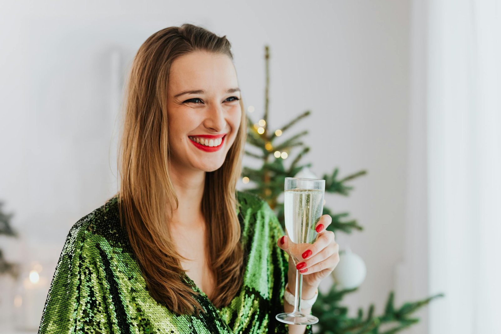 Smiling woman in green dress holding champagne glass by Christmas tree indoors.