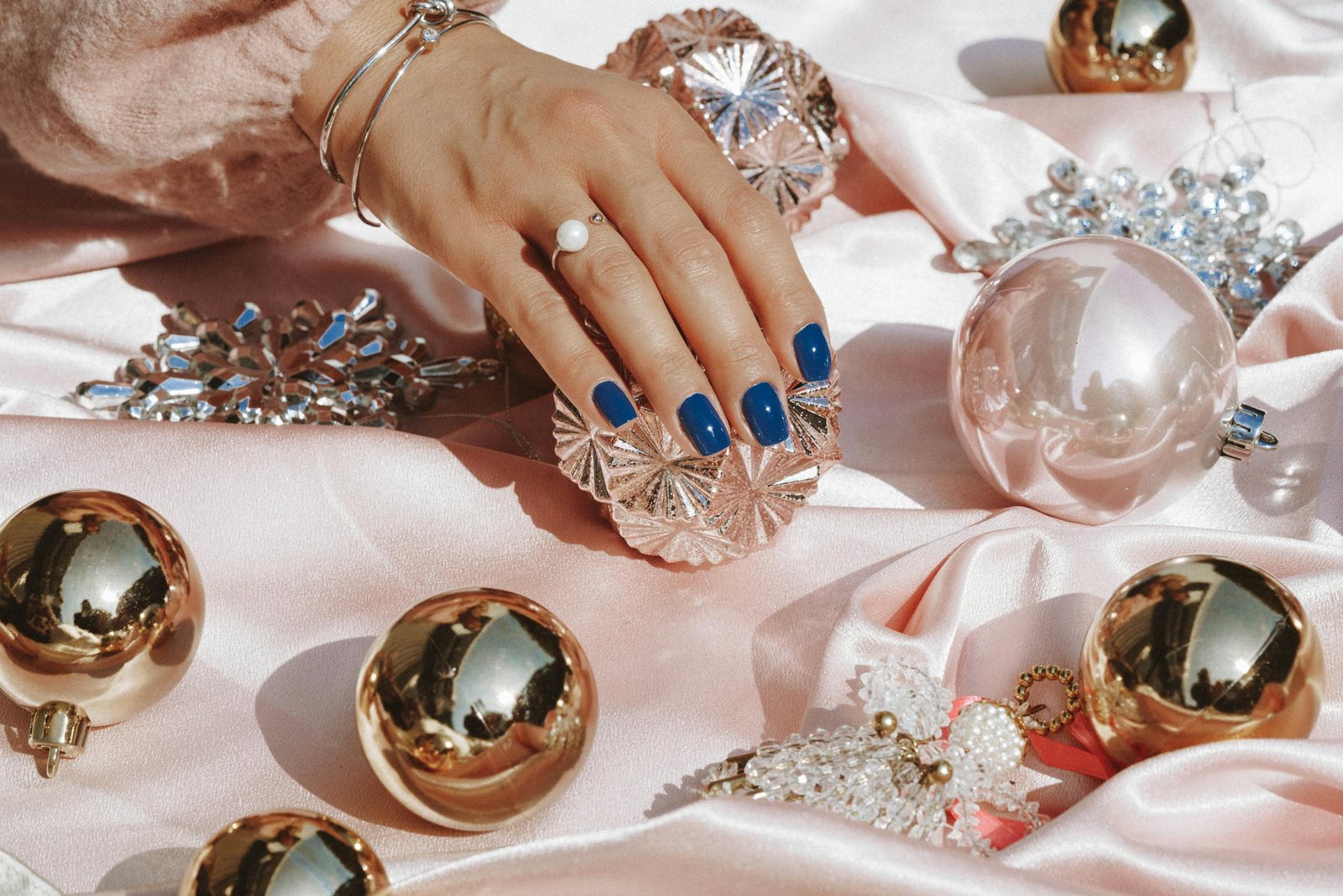 Stylish hand showcasing blue nails resting on pink satin surrounded by opulent ornaments and jewelry.