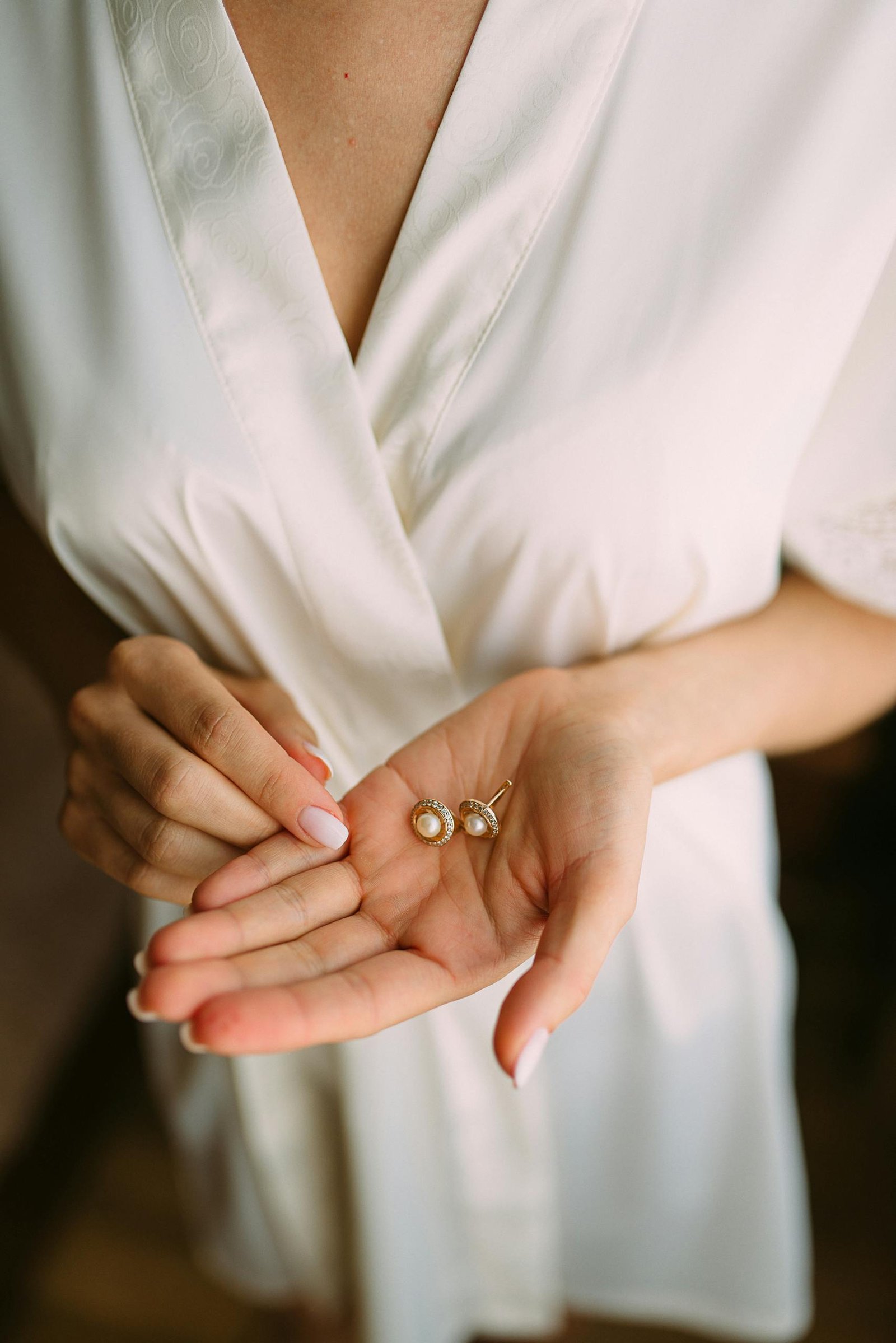 Close-up of a woman in white holding elegant pearl stud earrings, showcasing delicate hand and fashion detail.