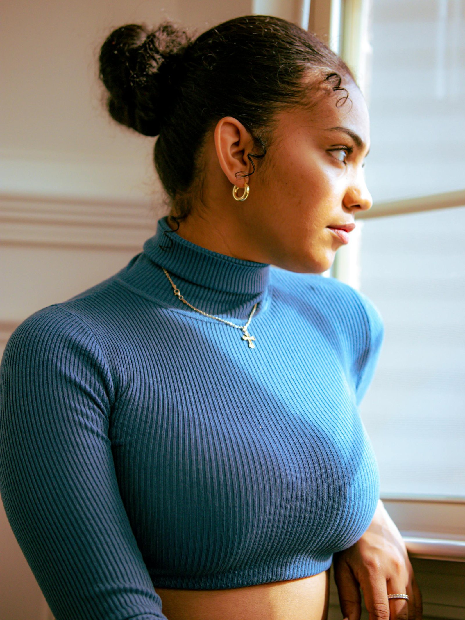 Portrait of a woman in a blue turtleneck gazing out a window, exuding elegance.