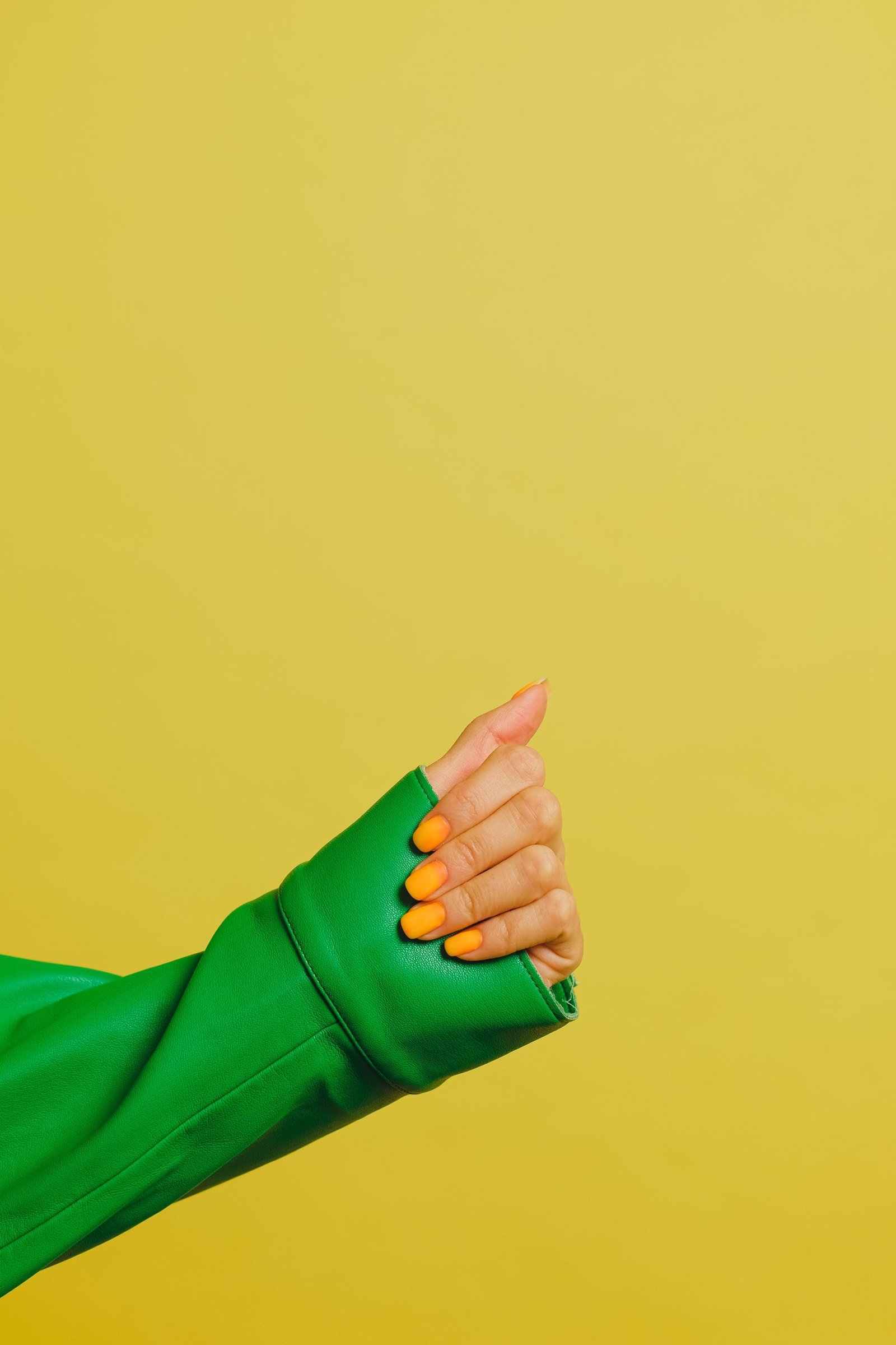 Close-up of a hand with orange nails and green jacket, vibrant yellow background.