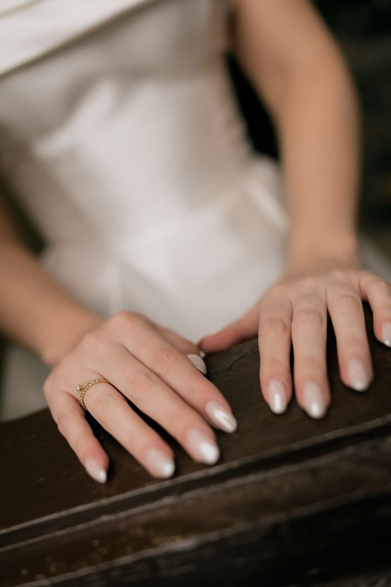 Elegant woman's hands with pearl nails and rings resting on dark wood, exuding sophistication.