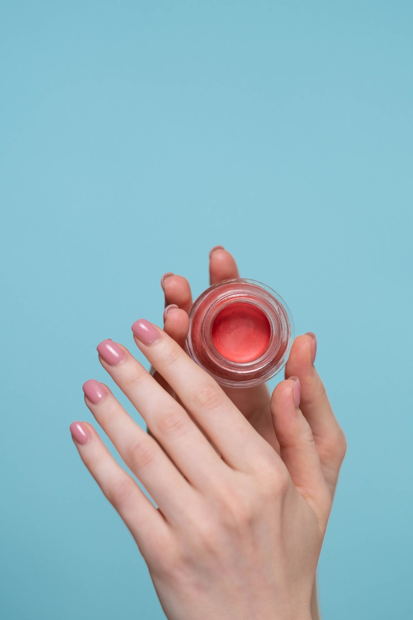 Minimalist photo of hands with pink nails holding a blush jar against a blue background.